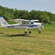 A white monoplane with a single engine flies just above the ground. The registration number "N15HV" is written in black near the rear of the fuselage.