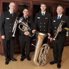 A set of four members of the U.S. Navy stand together in uniform, holding their respective brass instruments.