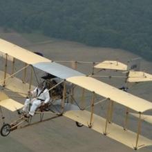 Diagonal side view of a biplane with yellow wings and a wooden frame during flight.