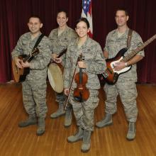 A group of four people pose together in U.S. Air Force camouflage uniforms. Each is holding an instrument: Two hold guitars, one plays a banjo, and the fourth plays the violin.