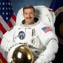 Dan Burbank, a male astronaut, stands for a photo wearing astronaut gear. He is standing in front of a background featuring a U.S. red, white, and blue flag and a second flag featuring NASA's seal.