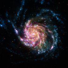 A still image of a spiral galaxy similar to the Milky Way galaxy, but larger in size. This galaxy features blue, white, pink, red, and yellow colors.