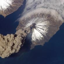 View from space of a volcanic eruption. A gray cloud of ash seen in the bottom left corner is floating away from the volcano site.