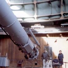 Three people pose near the bottom end of a cylinder-shaped object inside an observatory dome.