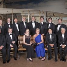 A large group of people sit and stand for a group picture after receiving a trophy from the museum. Two members sitting in the center hold the trophy.