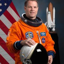 Commander Kevin Ford, a white male astronaut, poses for his official astronaut portrait in an orange suit. He is holding an astronaut helmet.