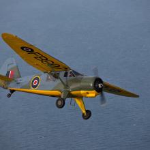 Side view of yellow, olive drab green, and gray monoplane in flight with one engine and fixed landing gear.