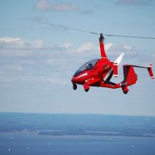 Side view of a small red helicopter in flight with one two-blade propellor above the cockpit.