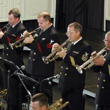 A group of musicians in U.S. Navy uniforms perform using various brass instruments.