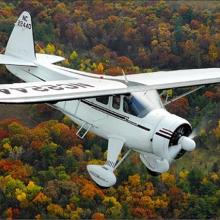 Diagonal front view of a white monoplane in flight with fixed landing gear and one engine.