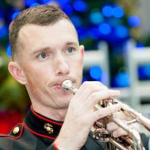 A man plays a trumpet in Marine Corps uniform as part of a larger group.