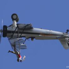 Side view of white and black biplane with single engine flying upside down. A person is depicted as standing on the upper wing of the plane.