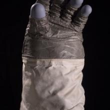 Astronaut glove featuring multiple different fabrics: a leather like fabric for the palm, a purple-colored rubber for the finger tips, and a tan fabric for the sleeve.