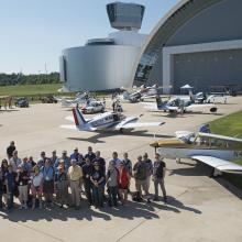 A group of individuals stand on a runway with several planes parked behind them.