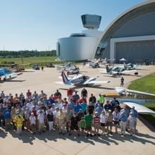 A group of pilots and museum staff pose as a group on a runway with planes behind them.