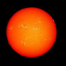 Disk view of the Sun's atmosphere revealing dark spots called sunspots and surrounding phages of lighter-colored clouds across the visible disk. Prominences of gases floating above the surface on the edges of the disk are also visible across the visible image.