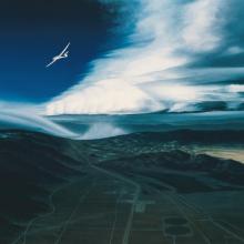 Painting of a arid lanscape with an airplane flying above clouds.