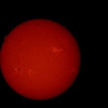 Disk view of the Sun's atmosphere featuring some sunspots (dark spots on the sun) surrounded by lighter-colored phages and a solar flare. A prominence and filament are visible as floating red gases on the upper left edge and a dark line on the upper center of the Sun, respectively.