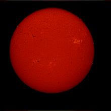 Disk view of the Sun's atmosphere with sunspot clusters (dark spots) surrounded by phages of lighter-colored clouds. A dark line known as a filament is also visible on the top left quadrant of the disk.