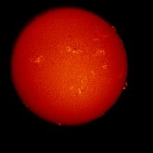 Disk view of the Sun's atmosphere. Dark spots known as sunspots are surrounded by lighter-colored clouds known as phages across the disk. Prominences, or red gases floating off the sun, are visible on the edges of the disk, mostly in the right half of the disk.