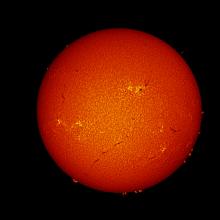 Disk view of the Sun's atmosphere. A large sunspot group is visible in the upper right quadrant as a large dark spot surrounded by lighter-colored phages. Other phages, darker lines called filaments, and floating gases from the edge of the disk called filaments are visible primarily on the bottom half of the disk.