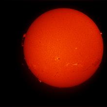 Disk view of the Sun's atmosphere. Several prominences, or red puffs of gases rising above the atmosphere of the Sun on the edge of the disk, are visible throughout the disk. Two sunspot clusters are visible with dark spots surrounded by lighter-colored phages on the bottom half of the disk.