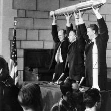 Three people hold a small, pencil-shaped satellite.