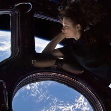 Tracy Caldwell Dyson, a white female astronaut, leans on a window showing the Earth in space.