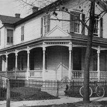 A large white house with a wrap around porch. A bicycle is leaned up against a short iron fence which surrounds the yard.