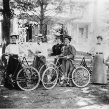 A group of five women in 1900s style dress with bicycles. 