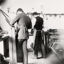 Two men with their back turned to the camera, working at a work bench and surrounded by tools.