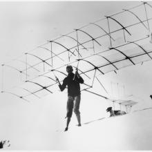 A man attached to a simple glider with two dogs behind him and a man holding up the glider on the right side.