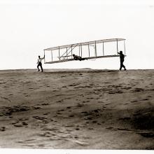 Orville Wright is prepared for takeoff in a test using the 1902 Wright Glider. The Wright Glider is being carried by Wilbur Wright and Dan Tate.