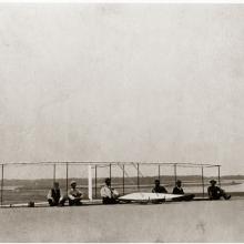 Six men sit on the ground in front of a glider