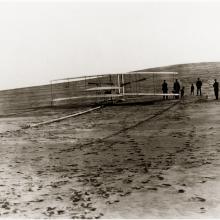 A biplane sits on a long wooden rail on the ground. A group of people stand behind it.