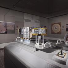 Multiple parts of the Rover robotic unit inside a display celebrating the Rovers. An electronic module made of multiple metal pieces can be seen on the left. On the right side, a cylindrical prism with one open side represents another part of the Rover.