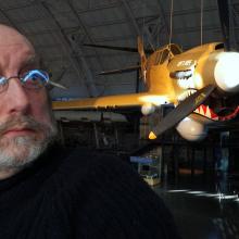 A white male specialist at the museum poses in front of a yellow monoplane with a shark mouth painted on the lower nose.