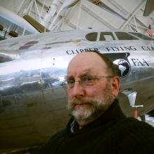 A white male archives specialist at the museum poses below a silver commercial airplane.