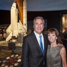 Two people, a white male and a white woman, pose together with a backdrop of a space shuttle model behind them.