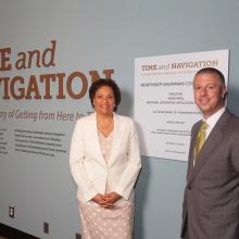 Two people, a woman and a male, stand next to the sponsorship plague for an exhibit that their corporation sponsored.