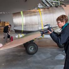 A camerawoman films a white male Museum specialist removing red tags from an aircraft.