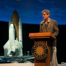 A white man who is a Curator of Space History at the Museum speaks at a podium. A backdrop includes a space shuttle.