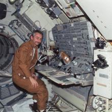 Owen Garriott, a male astronaut, stands near a console inside a space station.