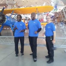 Three people who work as part of a puppetiering theater company stand together in front of a yellow and blue biplane inside the Museum's Steven F. Udvar-Hazy Center.