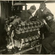 Three people examine a twelve-cylinder engine used for long-travel flight.