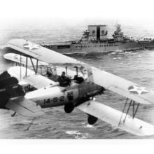 A military biplane flies over the ocean near an aircraft carrier.