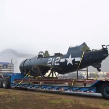 The fuselage of a blue military monoplane is transported on a blue truck between Museum facilities.