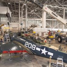 The engine of a blue military monoplane is placed by crane into the aircraft.