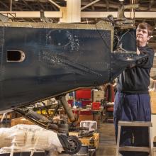 A museum specialist examines the tail and rudder of a blue military monoplane.