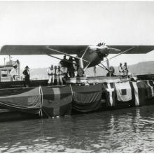 The Spirit of St. Louis sits on a barge as it is exhibited following Charles Lindbergh's record-breaking transcontinental flight. Flags are hung across the side of the barge.